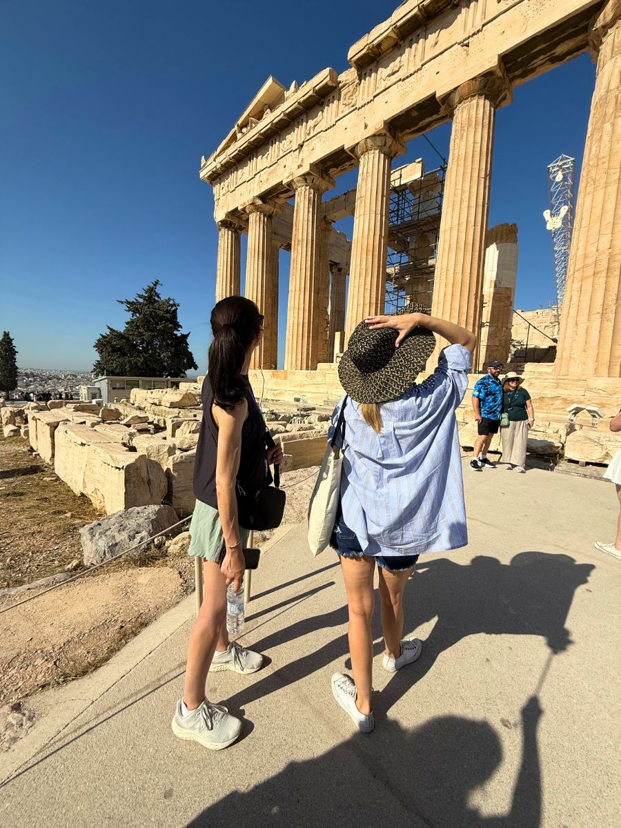 Two women admire the grandeur of the Parthenon in Athens under a bright blue sky, with ancient stone columns and scaffolding in view as they explore the historic Acropolis site.
