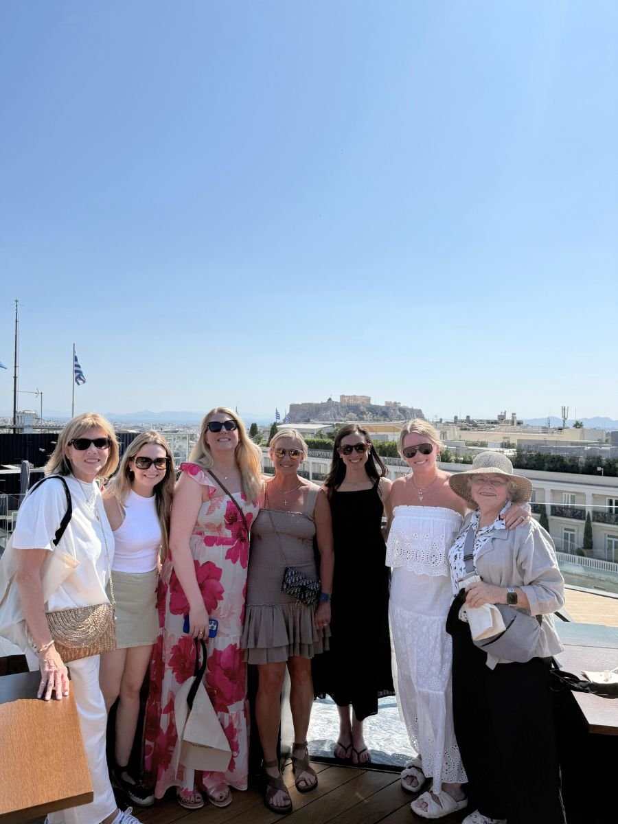 A group of seven women pose on a sunny rooftop with the Acropolis visible in the background. They’re dressed in light, summery outfits, smiling and enjoying the clear blue skies of Naxos, Greece.