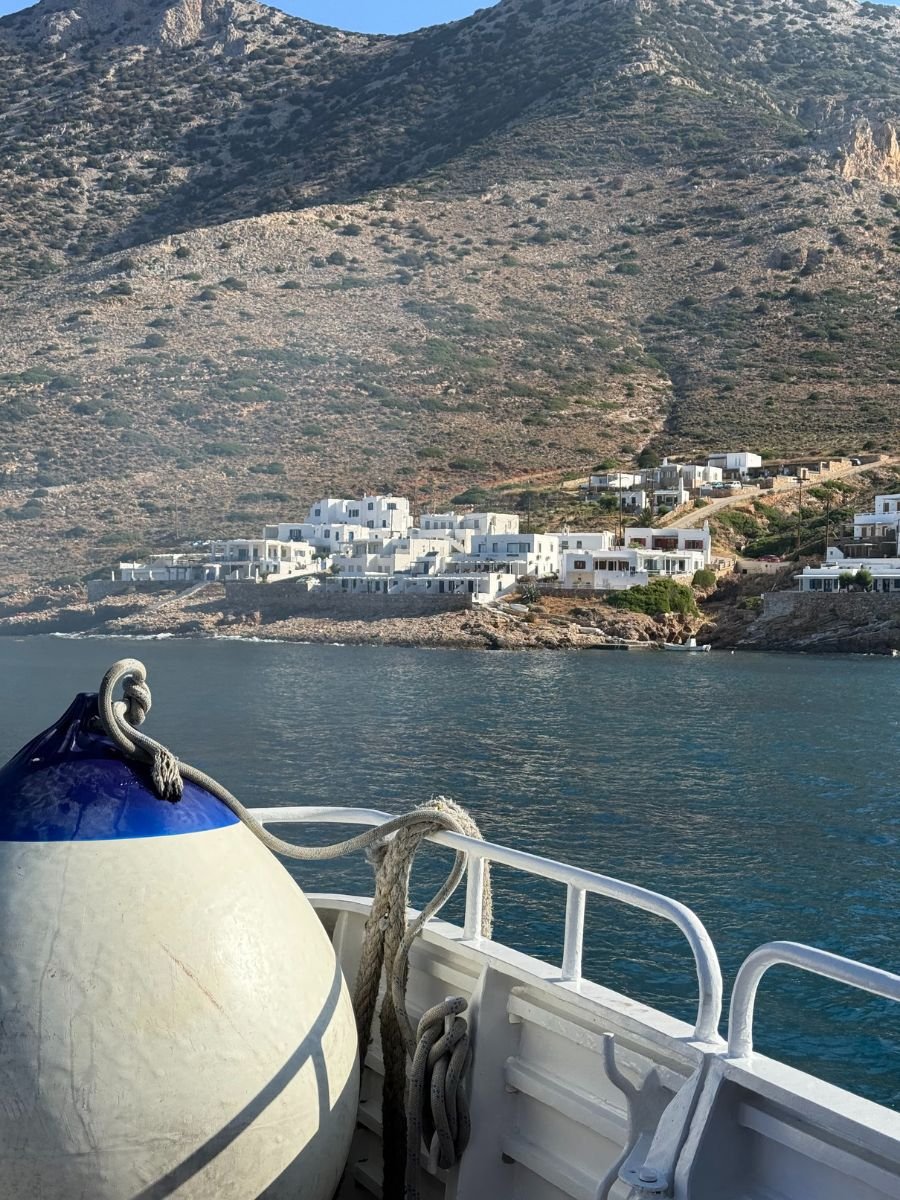View from a ferry deck showing a coastal village with white buildings built into a rocky hillside.