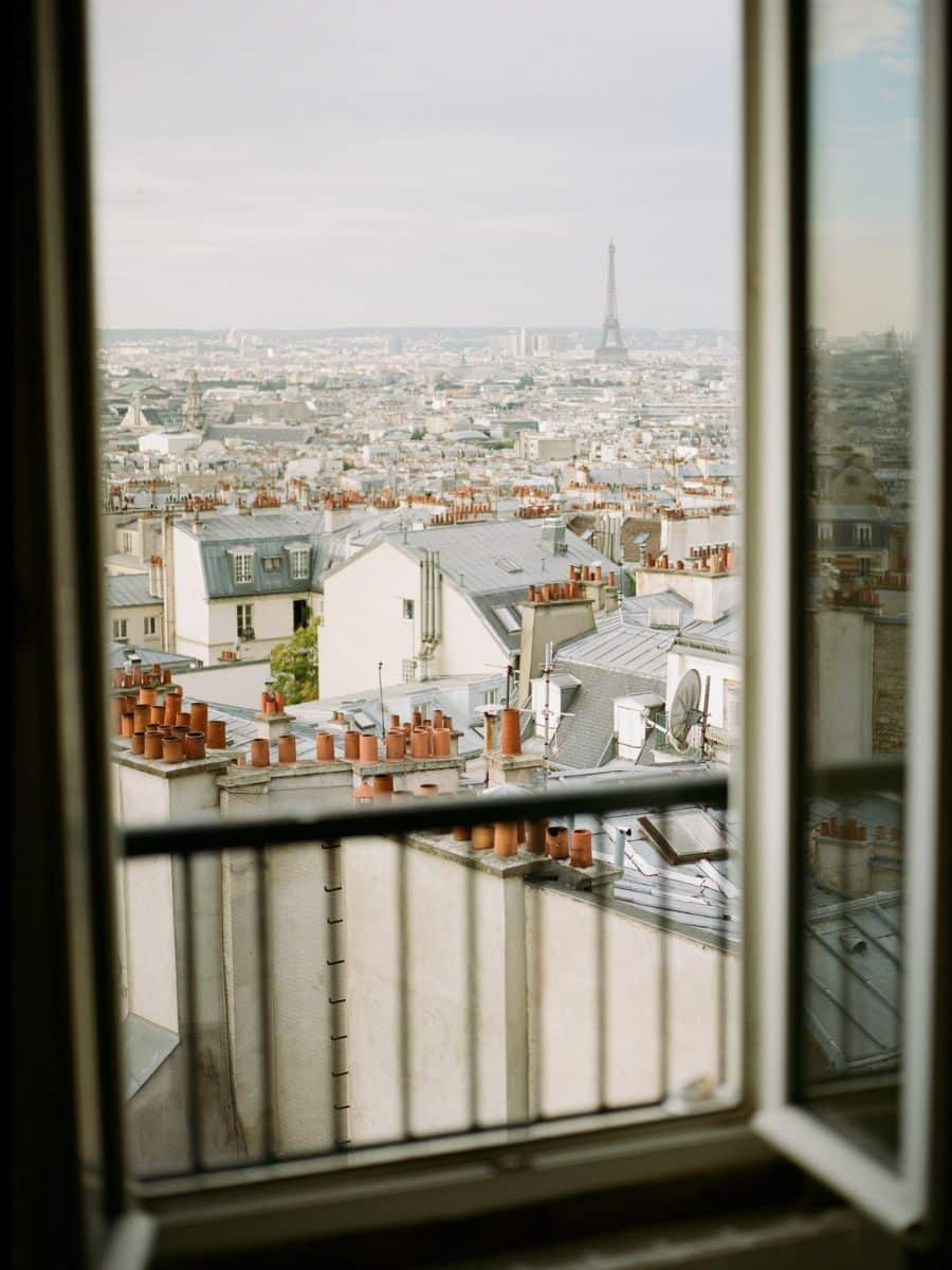 View of Paris rooftops through an open window, with terracotta chimney stacks and the Eiffel Tower in the distance under a soft, overcast sky.