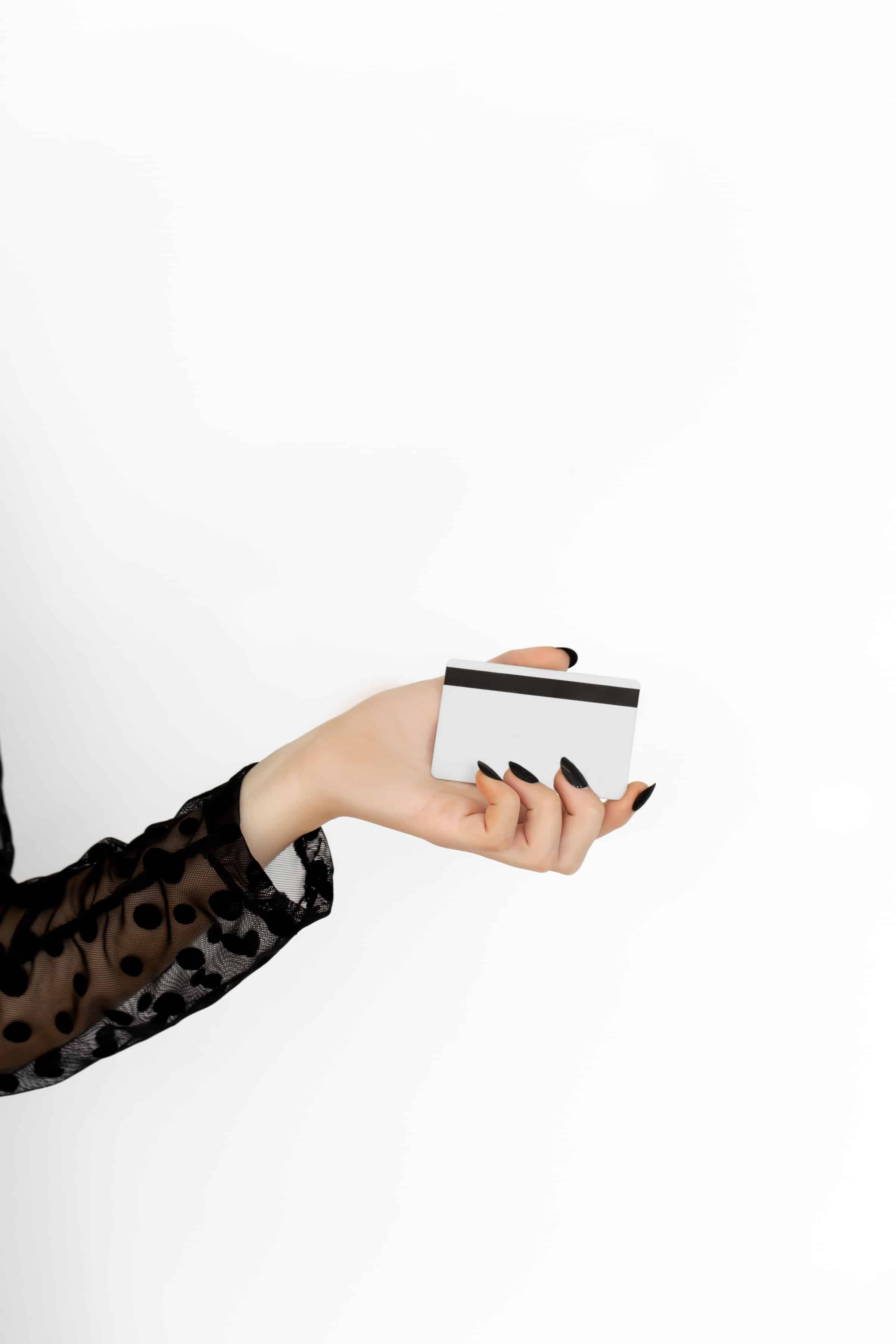 A close-up of a woman's hand with black nail polish holding a blank credit card, wearing a black sheer polka-dot sleeve against a white background.