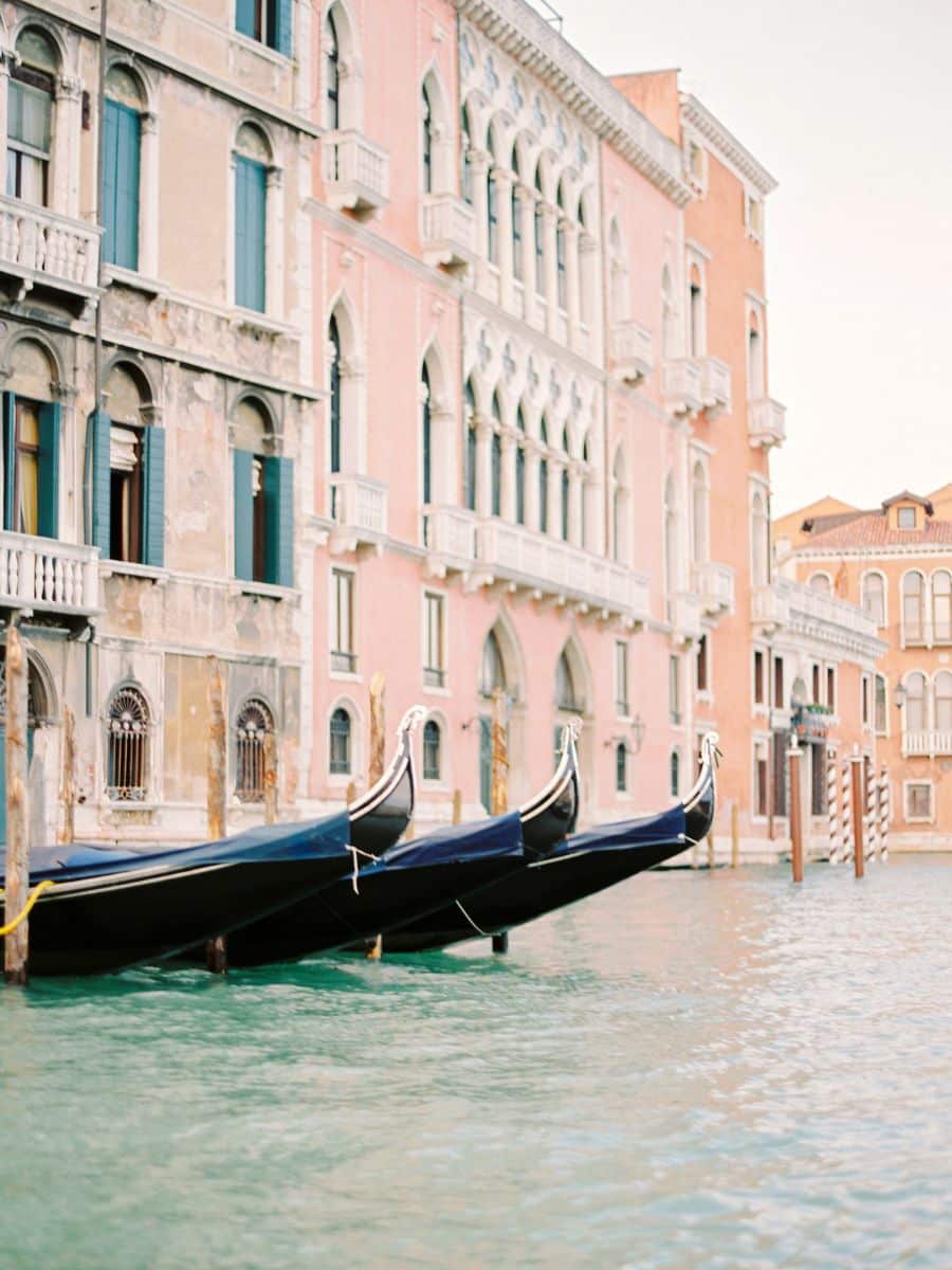 A row of gondolas gently bobbing on a canal in Venice, Italy, with elegant pastel-colored buildings featuring arched windows and balconies lining the waterway.