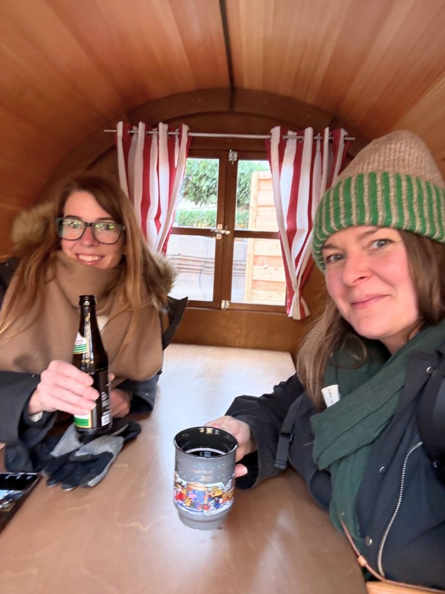 Two women inside a cozy wooden cabin, smiling and enjoying drinks. One is holding a Christmas-themed mug and the other a beer bottle, both bundled in winter clothes.