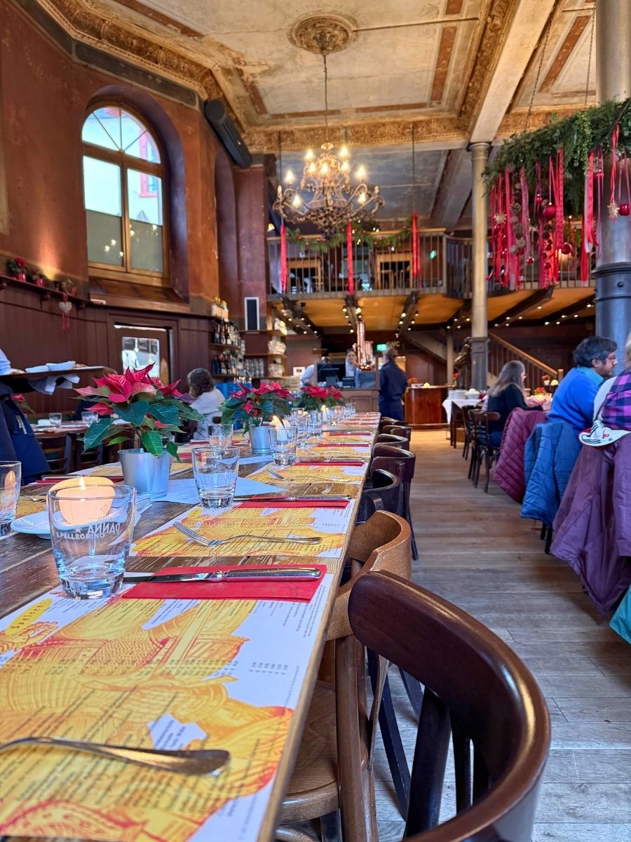 Long wooden dining table inside a grand historic restaurant, set with yellow map-themed placemats, red napkins, and poinsettias.