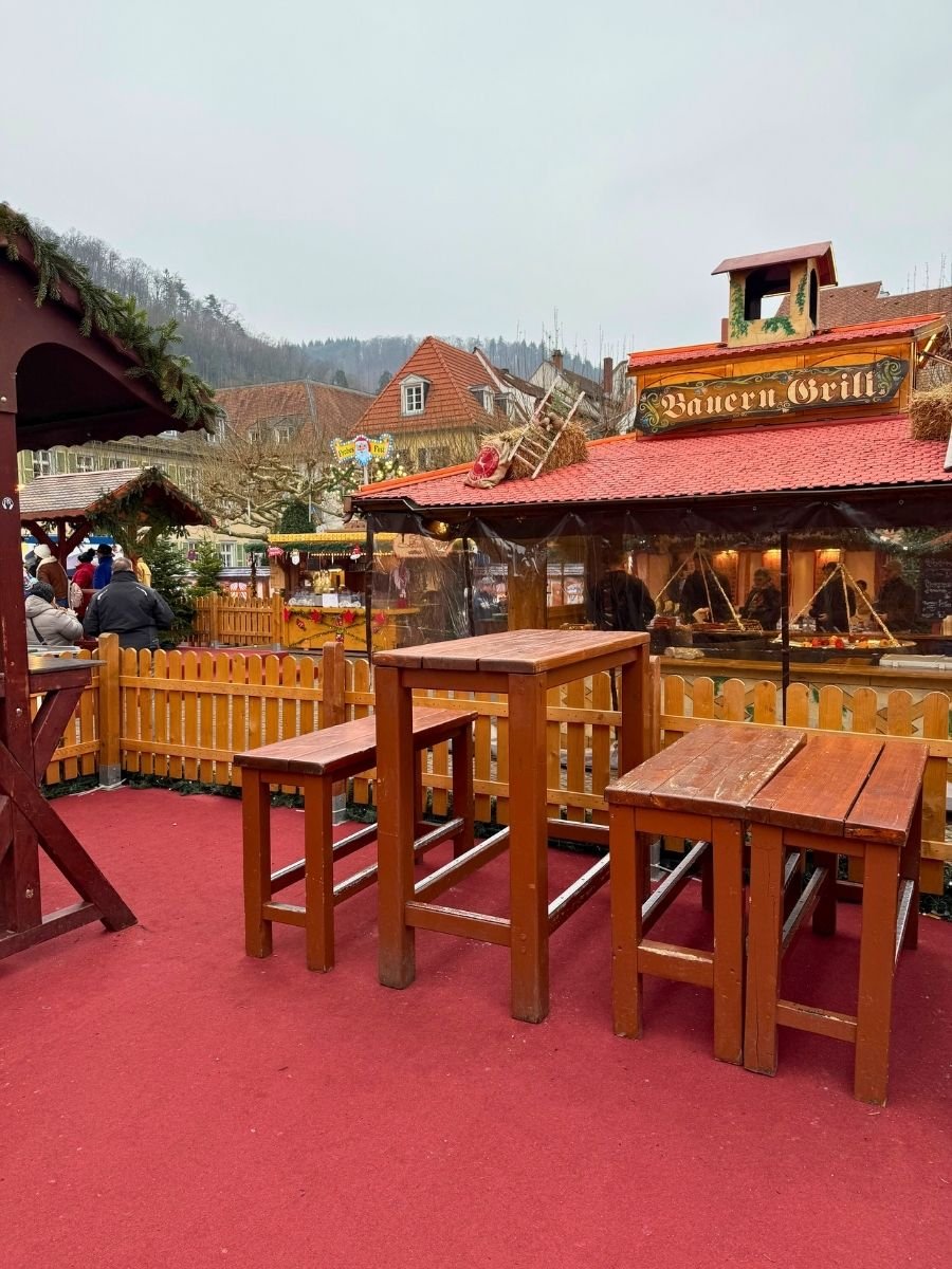 Outdoor seating area with tall wooden tables and stools at a Christmas market, with festive stalls and people browsing in the background.