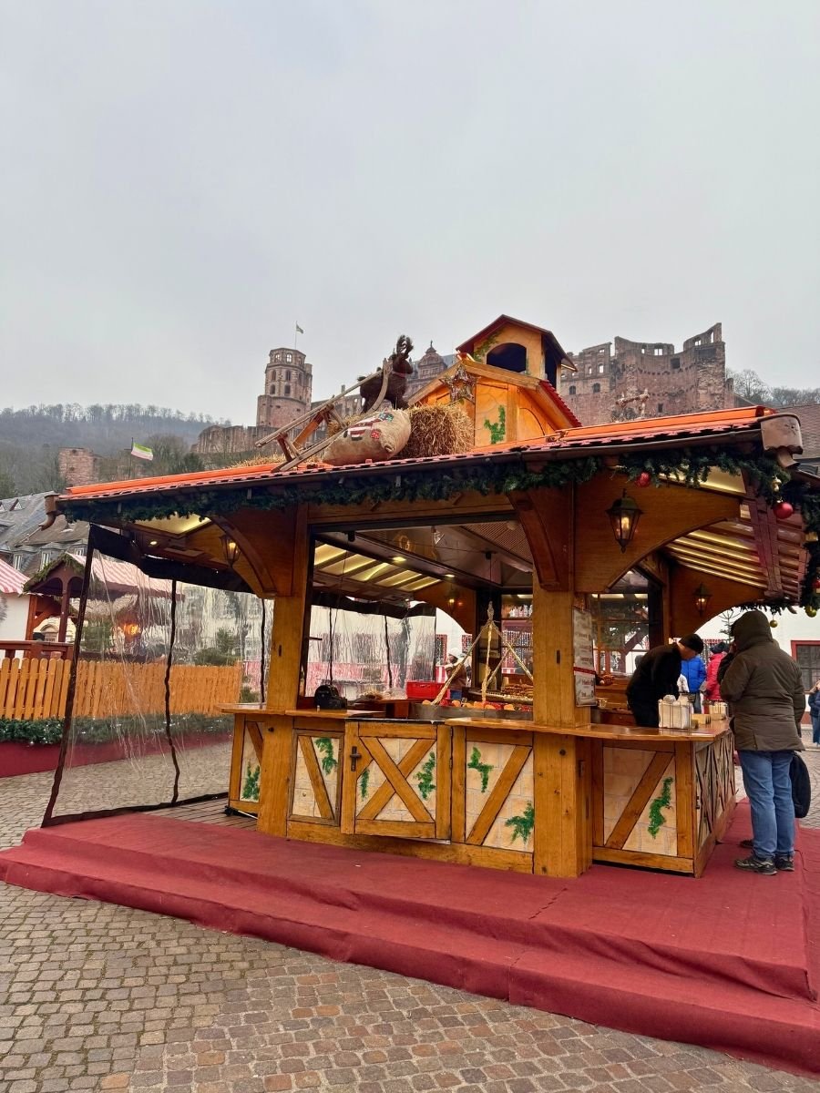Decorated wooden food stall at a Christmas market with Heidelberg Castle ruins visible in the foggy background.
