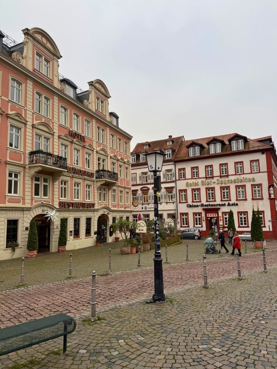 Charming cobblestone square in Heidelberg with pastel historic buildings, including Hotel Holländer Hof and a Chinese restaurant.