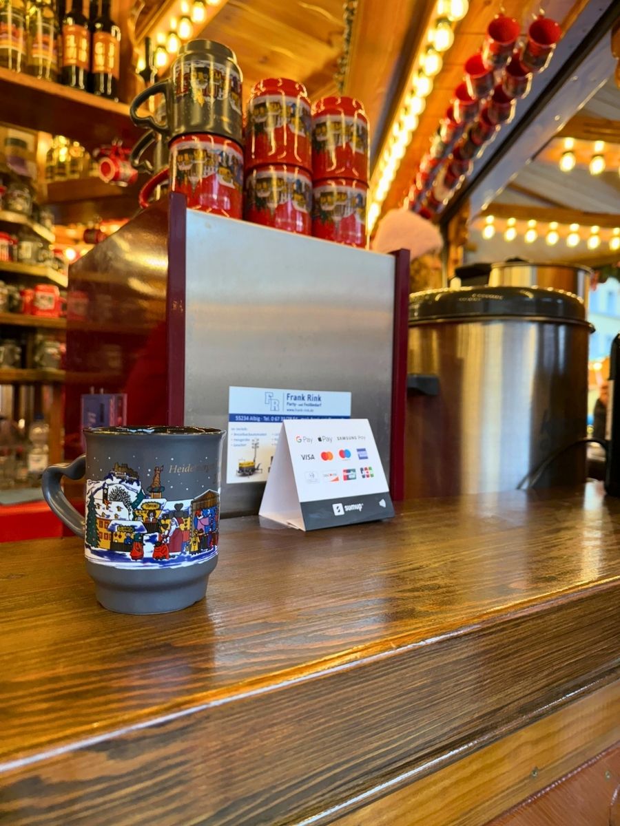 A festive wooden market stall decorated with lights and colorful souvenir mugs, featuring a ceramic mug with a winter village scene on a polished wooden counter.