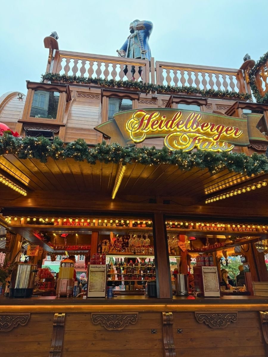 A decorated wooden bar stall at the Heidelberg Christmas Market, with a sign reading “Heidelberger Fass” and a statue of a man in historical clothing standing on top.