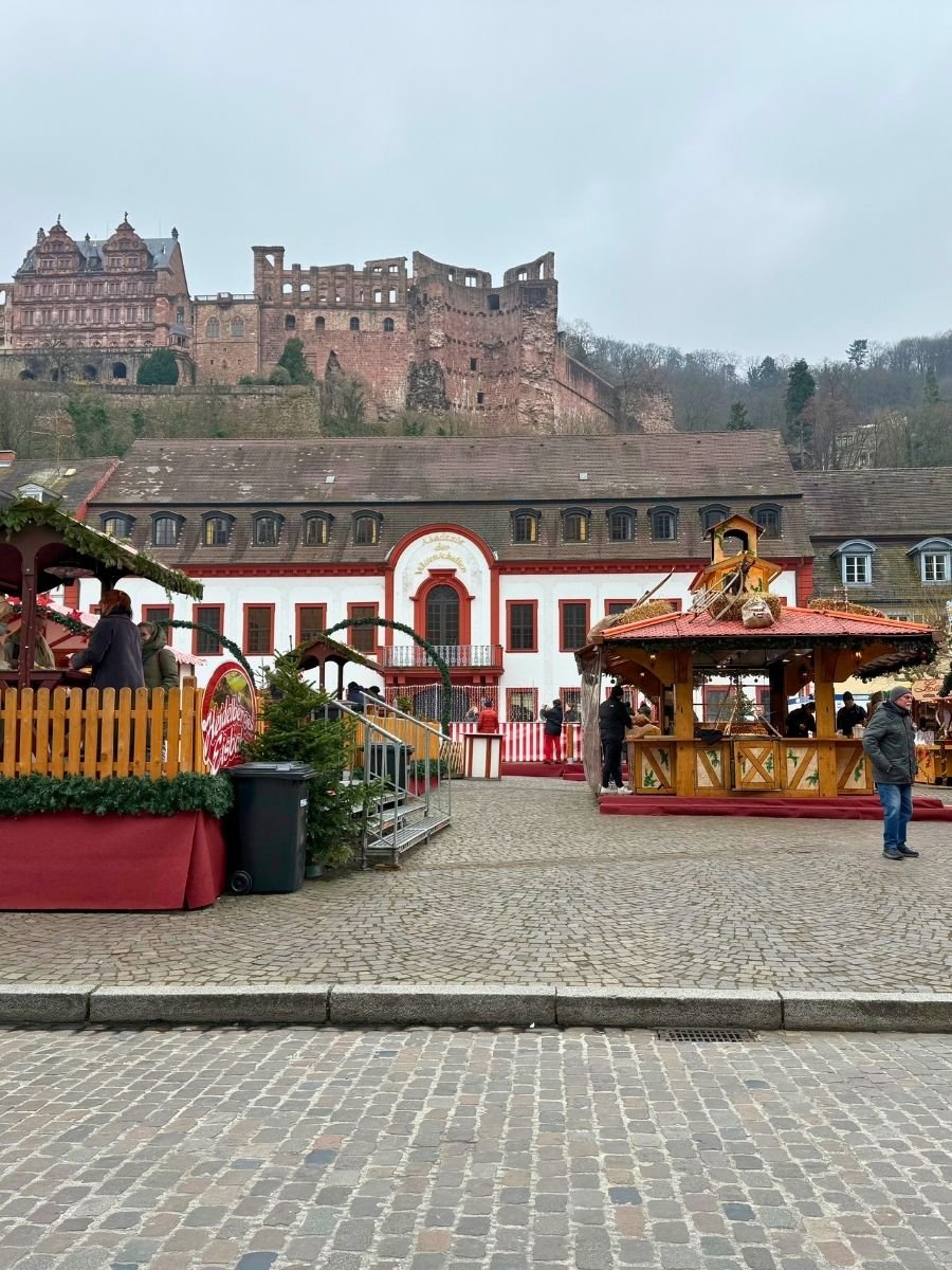 A festive scene at the Heidelberg Christmas Market with a view of the red-and-white building in front of the castle ruins, and cozy market huts lining the square.