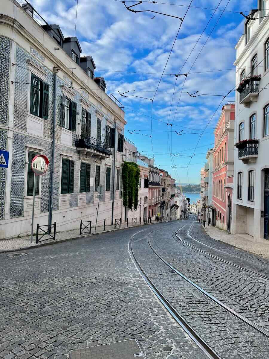 A steep, cobblestone street in Lisbon, lined with beautifully tiled buildings and balconies, with tram tracks running down the center. The street curves gently toward the horizon, where the bright blue sky and the Tagus River can be seen in the distance. Overhead, a network of tram cables crisscrosses against a backdrop of clouds.