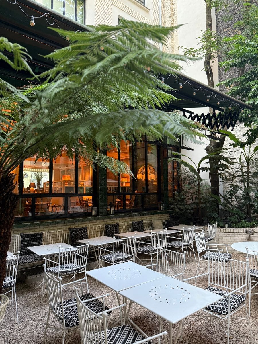Lush courtyard garden dining area at Hotel Eldorado Paris with white metal furniture and string lights under green foliage.