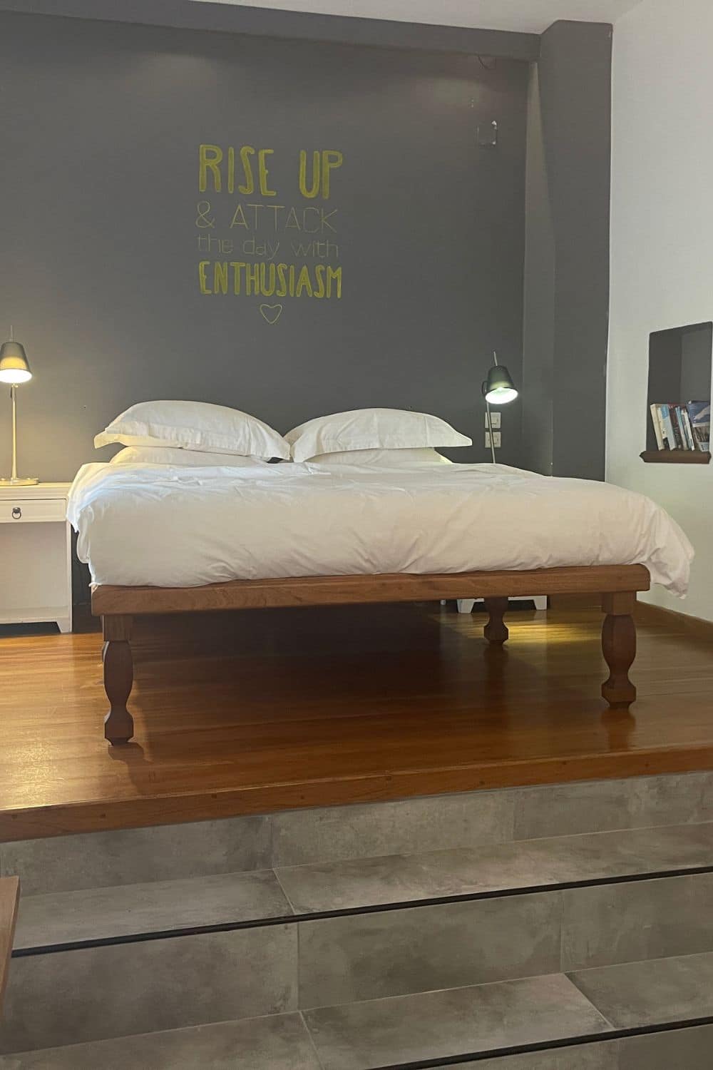 Minimalist hotel room in Hydra with a motivational quote 'RISE UP & ATTACK the day with ENTHUSIASM' on the dark grey wall above a wooden bed, inviting a positive start to the day.