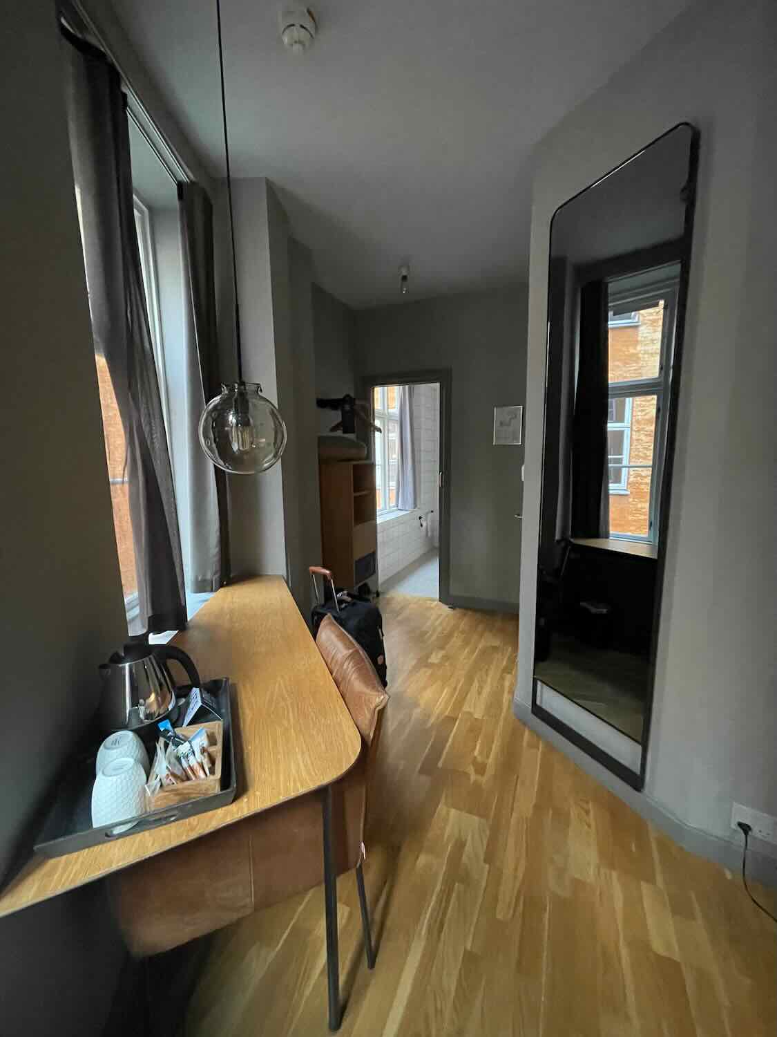 Interior shot of a room at Hotel SP 34 showing a sleek workspace with a curved wooden table, a floor-length mirror enhancing the space, and a peek into the adjacent bedroom area, showcasing the hotel's chic and functional design.