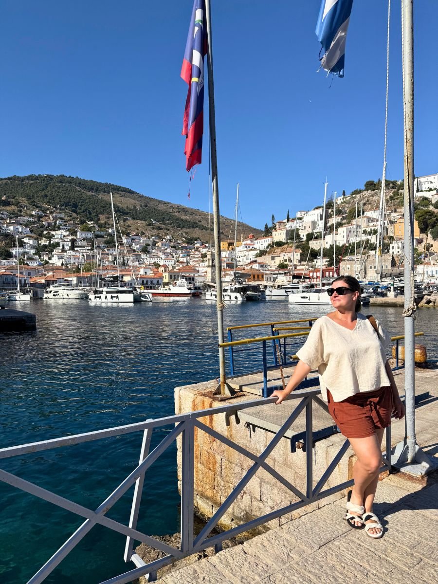 Melissa standing by the harbor railing in Hydra, Greece, with a scenic view of hillside whitewashed houses, docked sailboats, and flags waving under a clear blue sky.