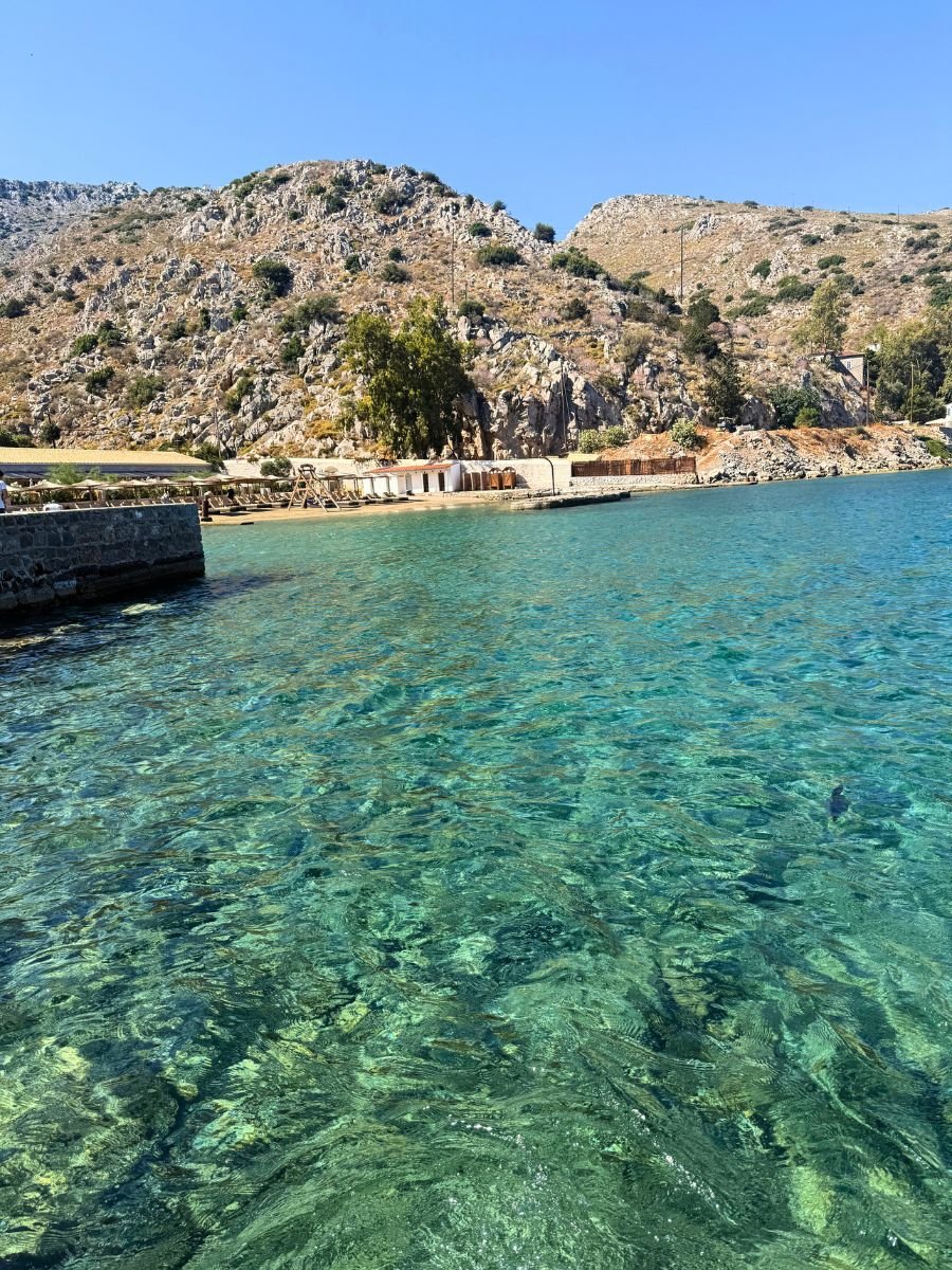 Crystal-clear turquoise waters of Hydra, Greece, with rocky hills and a small beach area in the background under a bright blue sky.