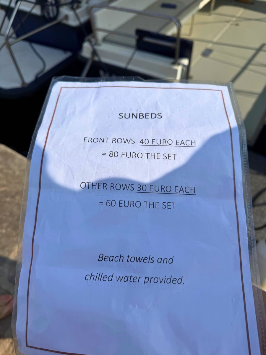 A printed sign listing sunbed rental prices in Hydra, Greece. It states that front row sunbeds cost 40 euros each (80 euros per set), and other rows are 30 euros each (60 euros per set). The sign also mentions that beach towels and chilled water are provided.