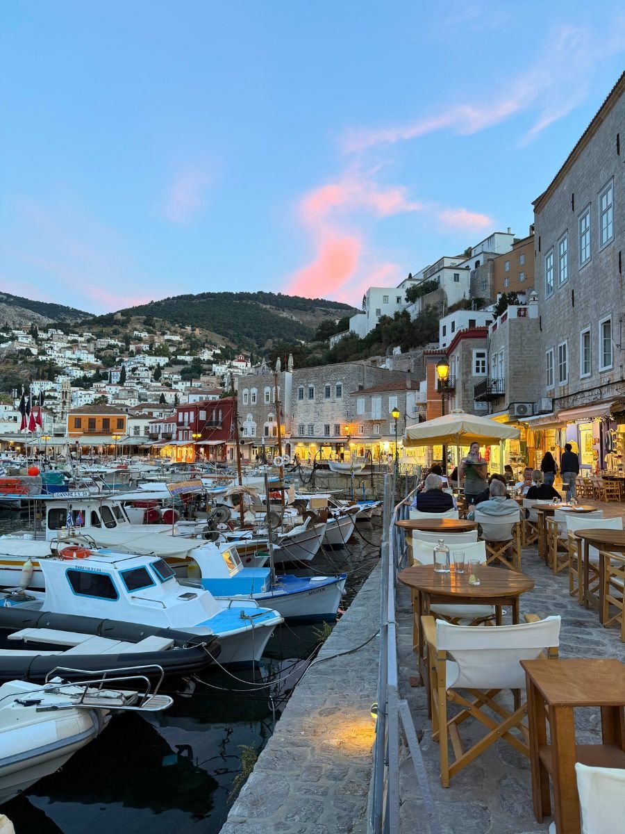 Evening view of the Hydra harbor in Greece, with docked boats, waterfront restaurants, and diners enjoying the sunset under a sky painted with soft pink clouds and blue hues.