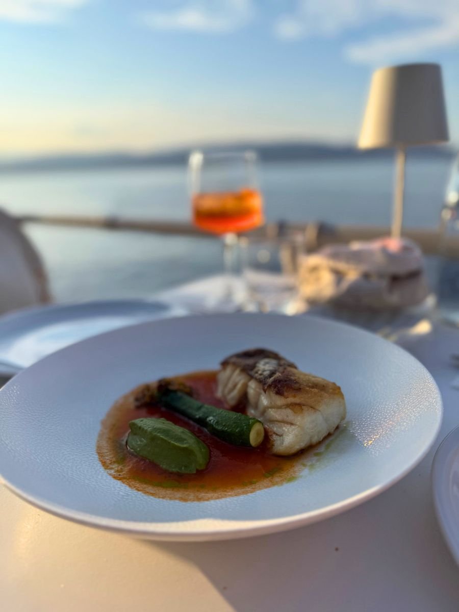 A gourmet fish dish served with zucchini and green puree in a tomato-based sauce, elegantly plated at a seaside restaurant in Hydra, Greece, with a sunset ocean view and an Aperol spritz in the background.