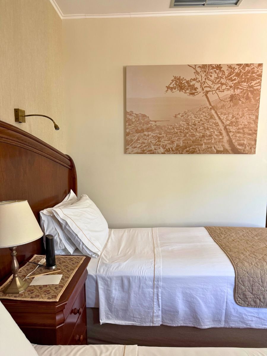 Cozy hotel room at Leto Hotel in Hydra, Greece, featuring twin beds with crisp white linens, a wooden headboard, a nightstand with a lamp, and a sepia-toned wall art depicting a scenic view of the island.
