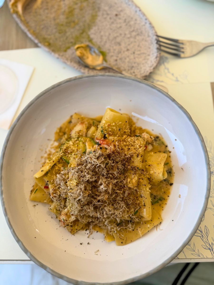 A bowl of wide pasta noodles topped with freshly grated truffle, served in a creamy sauce with herbs and visible seasoning. An empty appetizer plate and utensils are in the background.