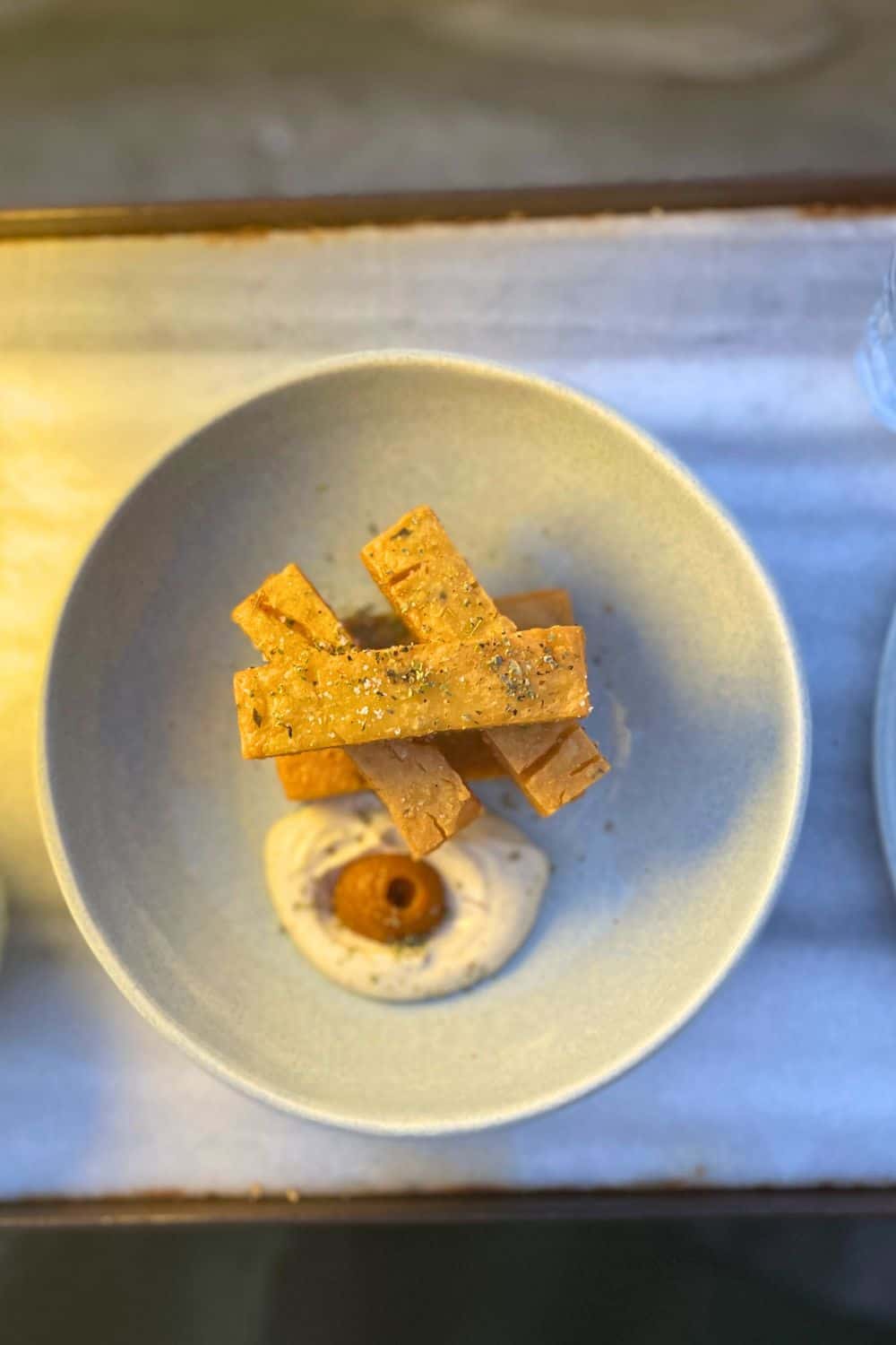 The image shows a minimalist dish served in a light-colored bowl. The dish consists of several rectangular pieces of crisp bread or crackers, arranged neatly on top of a creamy spread or dip. A small round component, possibly an olive or similar garnish, is placed at the center of the spread, giving the dish an elegant and simple presentation. The lighting and composition emphasize the refined, gourmet style of the meal.