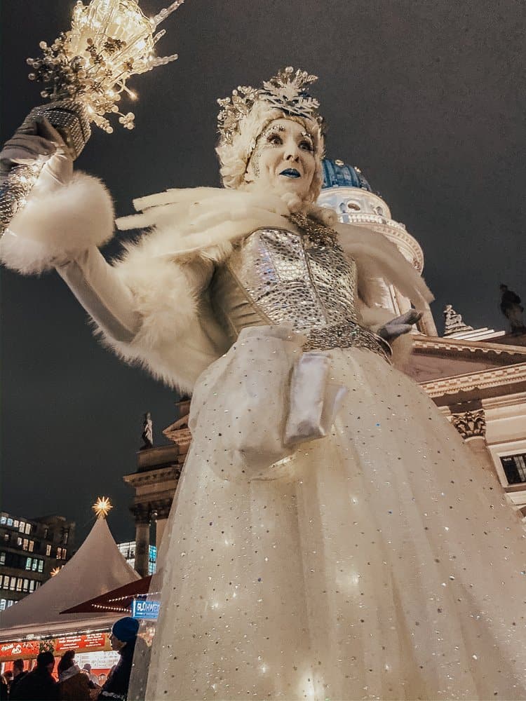 A close-up of a performer dressed as a winter queen in a sparkling, white gown with a fur-trimmed cape. She is holding a glowing staff and wearing an elaborate crown. The scene is set against a night sky with festive lights and a domed building in the background, enhancing the magical and regal atmosphere.