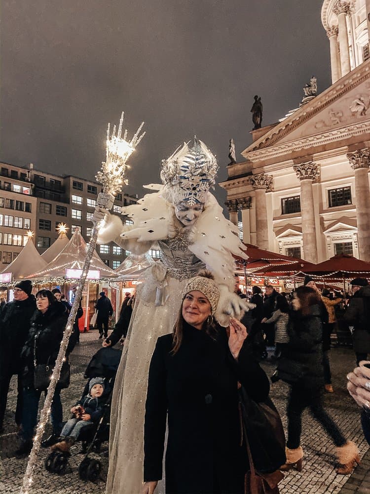 A festive scene at a Berlin Christmas market with a woman posing near a performer dressed in an elaborate white and silver winter-themed costume, towering on stilts.