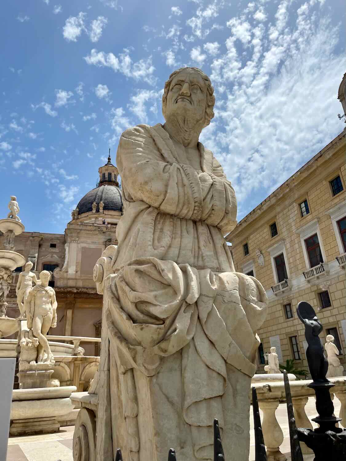 A statue of a robed figure is set against a backdrop of trees and a historic building with a dome in Catania, Sicily. The Italian flag waves in the background under a clear blue sky.