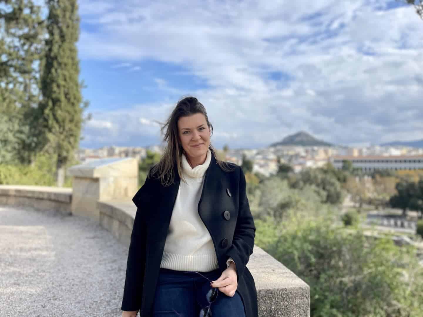 A confident solo traveler smiles gently, seated on a stone balustrade with a panoramic view of a sprawling cityscape under a cloudy sky. She's stylishly attired in a warm turtleneck and classic black coat, embracing the cool air, with the promise of adventure in her eyes.