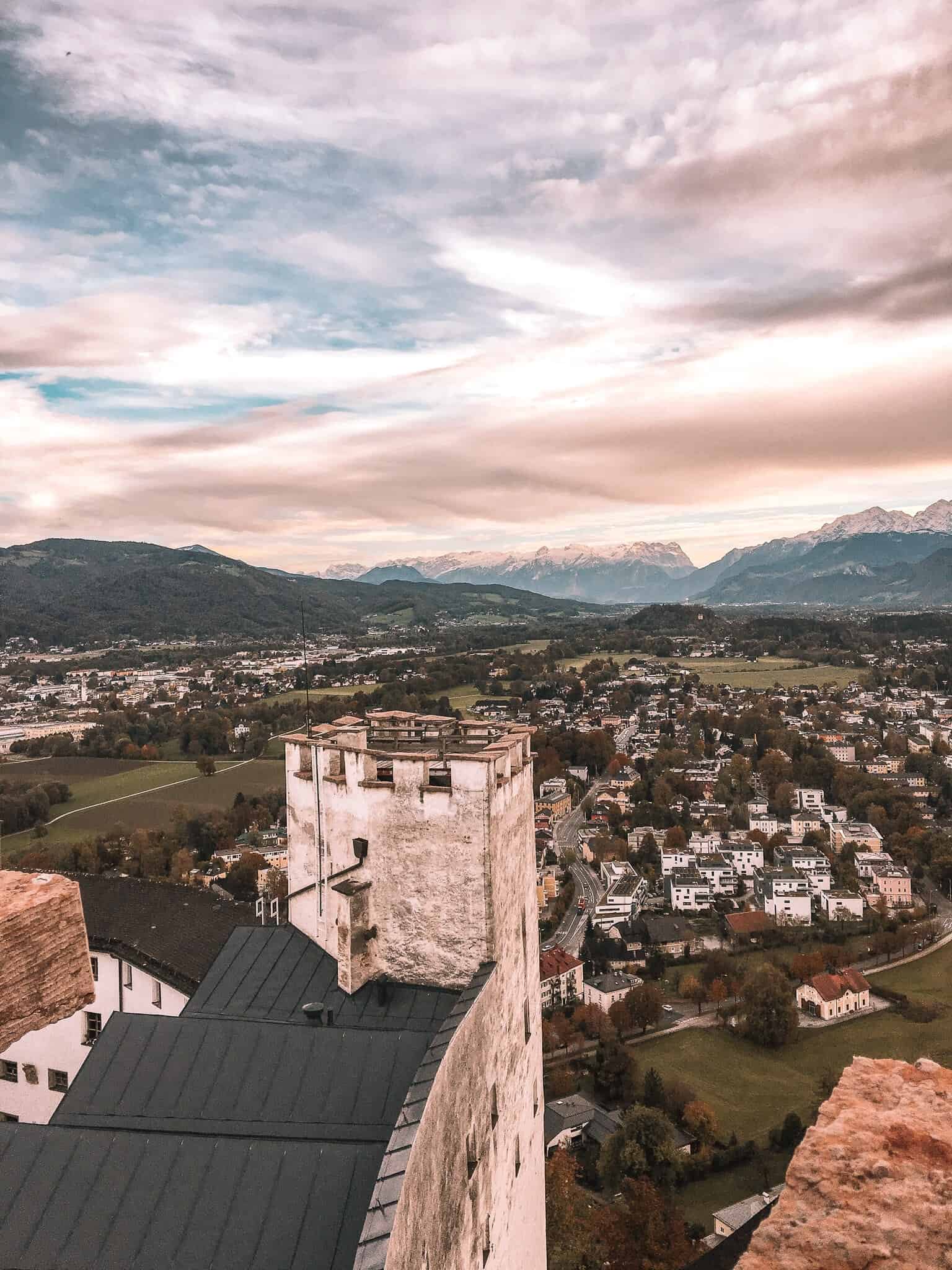 View from Hohensalzburg Fortress overlooking Salzburg, Austria, with the city’s rooftops, green fields, and distant mountains under a dramatic cloudy sky.