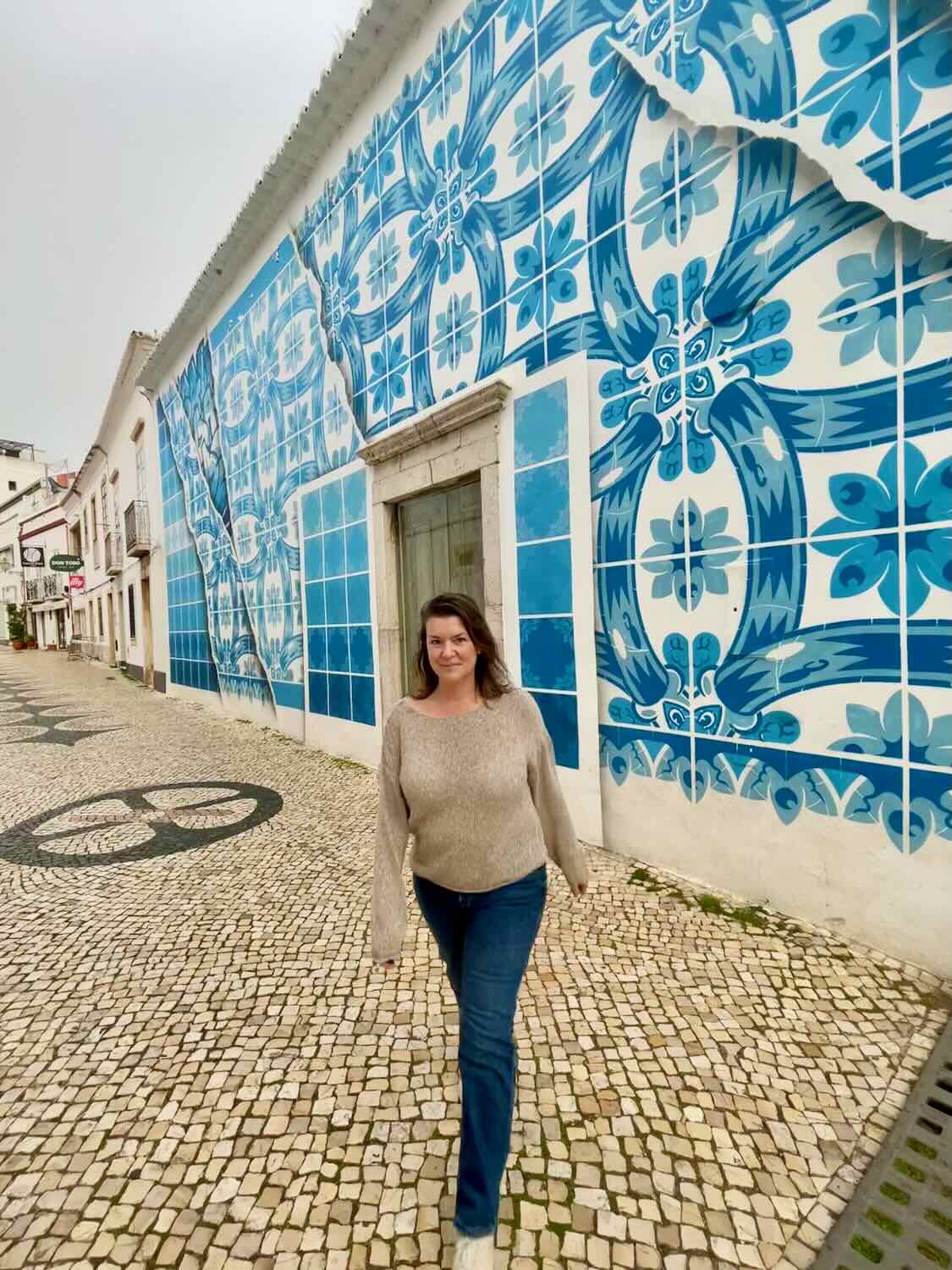 A woman walking on a cobblestone street in front of a building adorned with intricate blue and white tile designs in Portugal. She is wearing a beige sweater and blue jeans, with a relaxed and happy expression