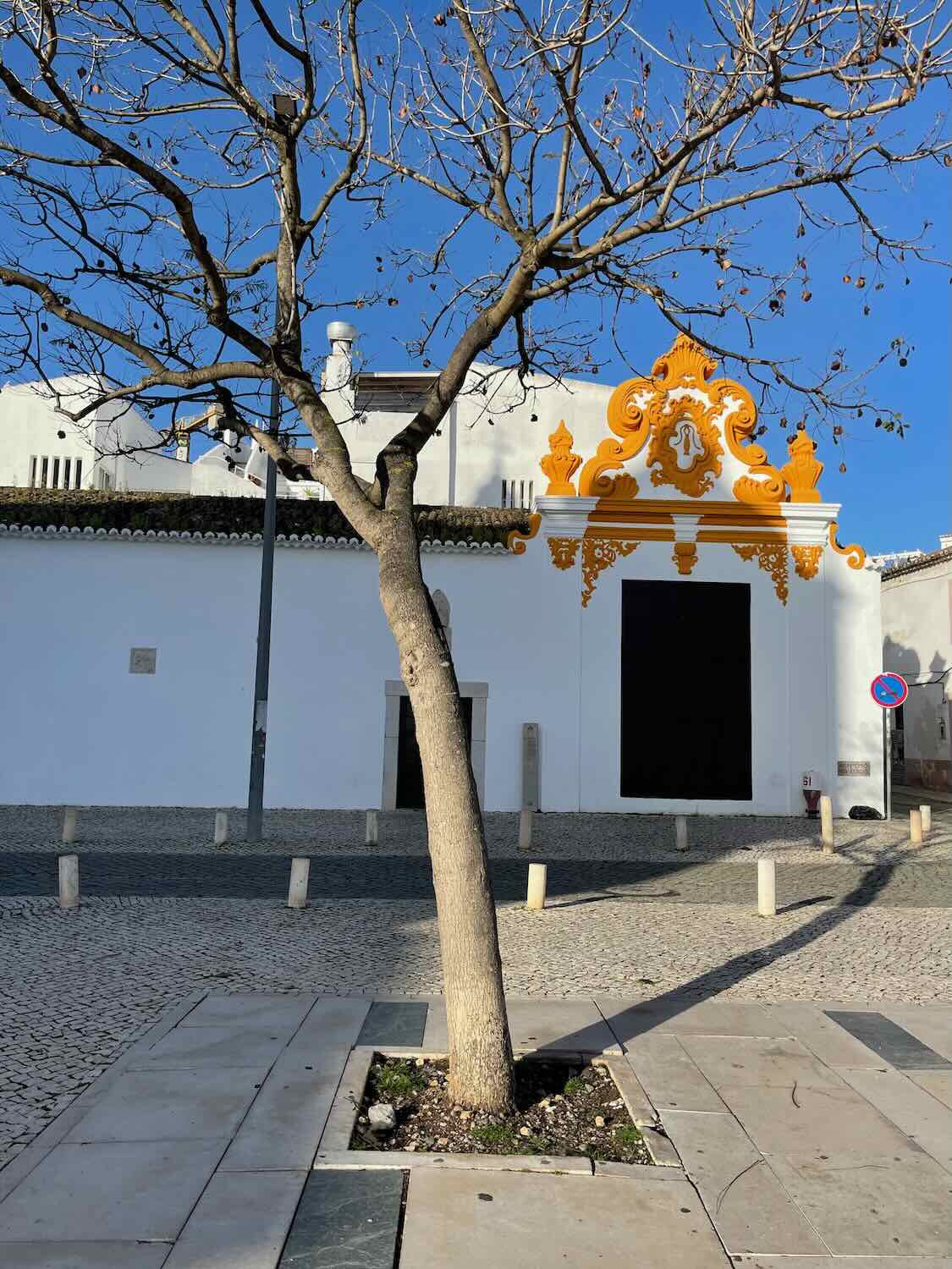 A leafless tree standing in front of a building with ornate orange and white architectural details on a sunny day. The scene is captured on a cobblestone street with a clear blue sky in the background.