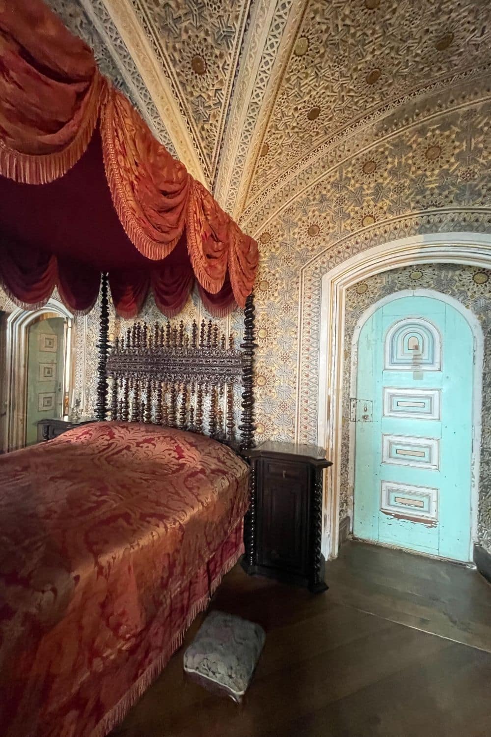 Ornate bedroom inside Pena Palace featuring a dark carved four-poster bed with red damask bedding and a dramatic canopy. The arched ceiling and walls are covered in intricate geometric patterns, and a weathered turquoise door adds a pop of color to the richly decorated room.