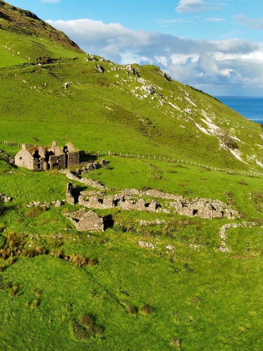A scenic view of lush green hills along Ireland’s coastline with the ruins of old stone cottages scattered across the landscape under a bright blue sky with scattered clouds.