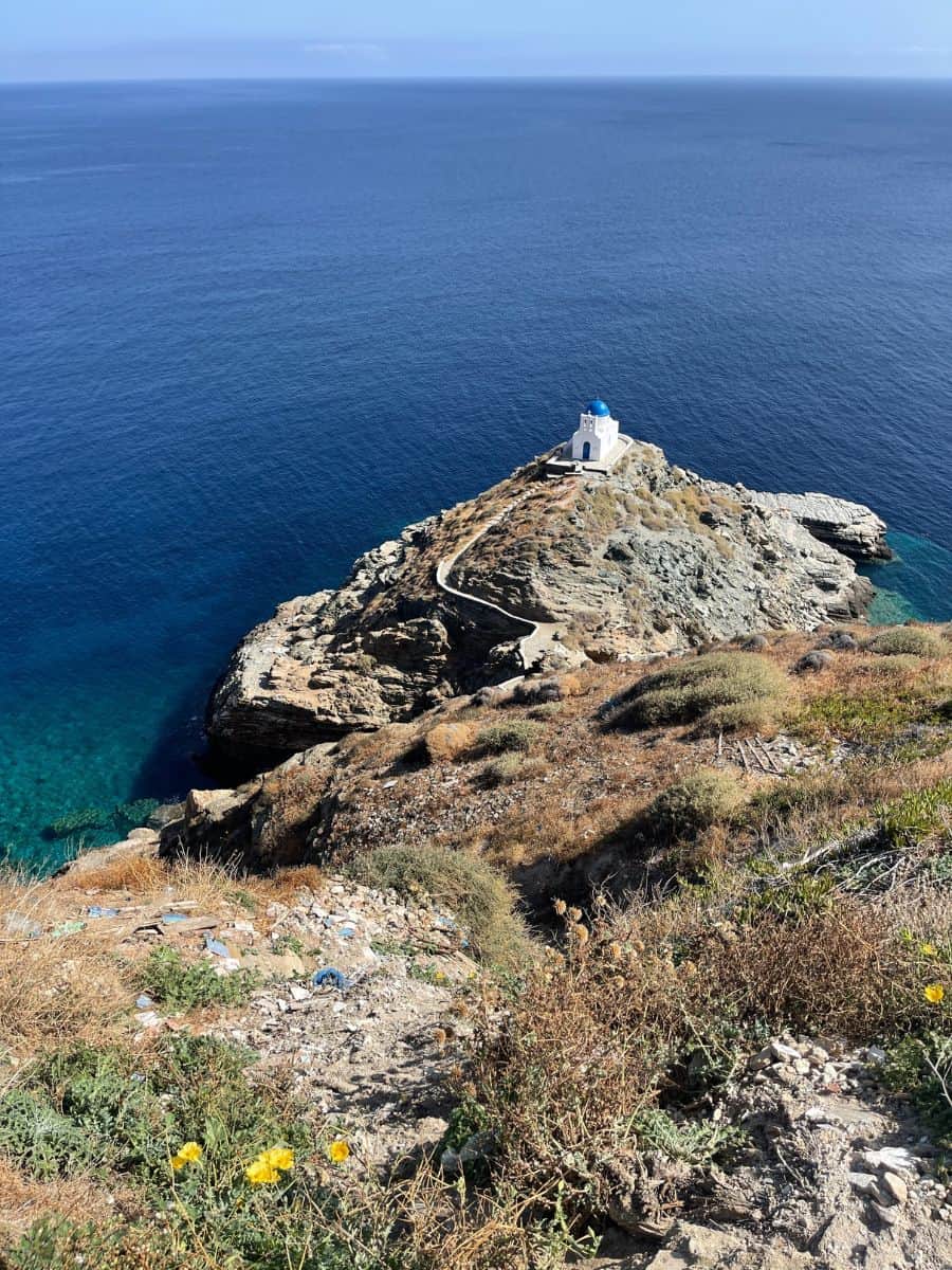 Scenic view of the Church of the Seven Martyrs perched on a rocky cliff in Sifnos, Greece, overlooking the deep blue Aegean Sea, with a winding path leading to the whitewashed chapel.