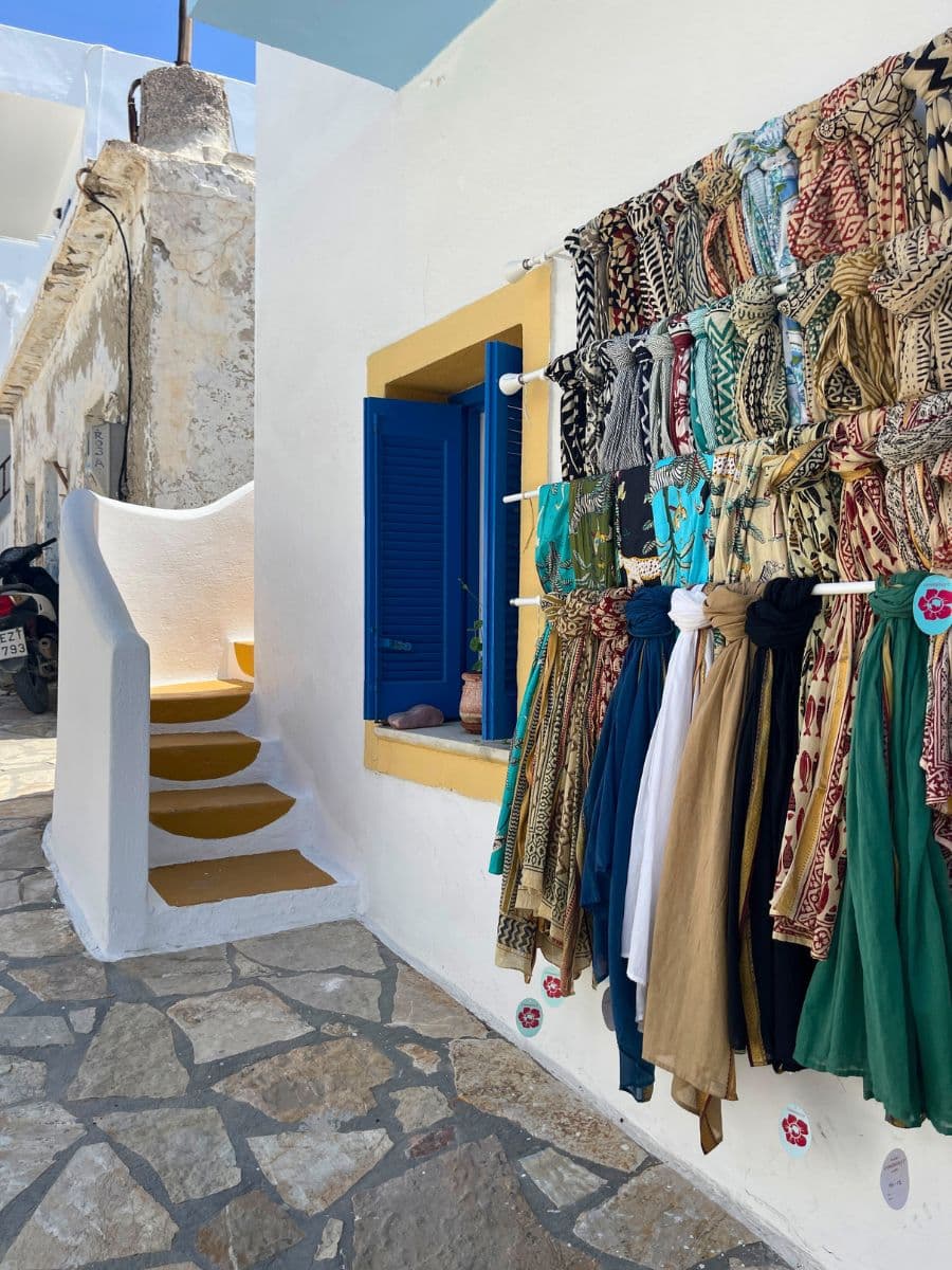 A boutique shop displaying colorful dresses and scarves, set against a traditional Greek whitewashed building with blue accents.
