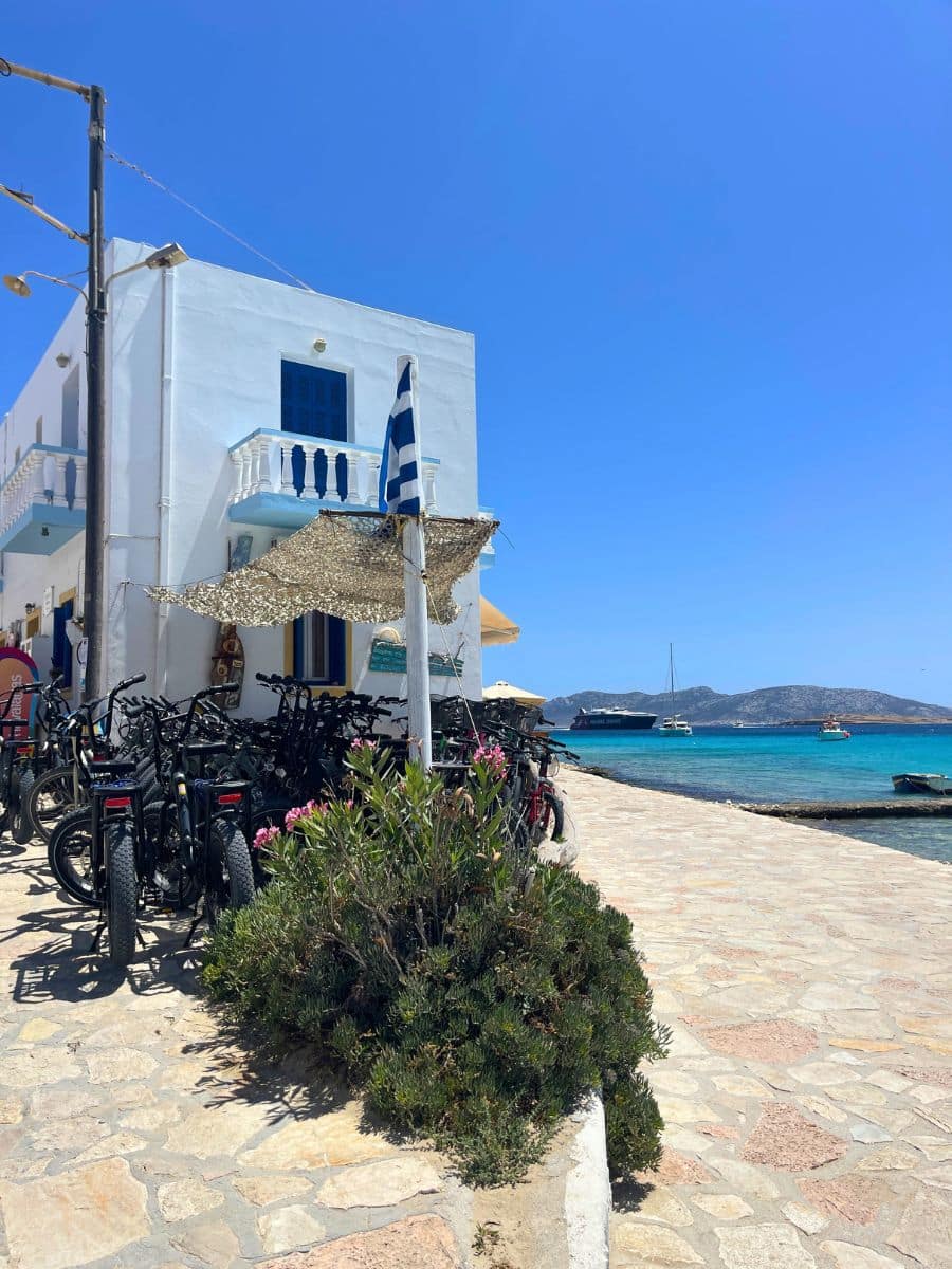A coastal scene with bicycles parked along a stone walkway, a white Greek building with blue shutters, and a Greek flag waving in the breeze.