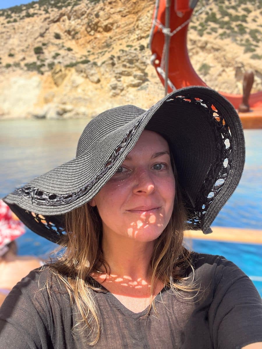 A woman wearing a wide-brimmed black hat takes a selfie on a boat, with clear blue waters and rocky cliffs in the background.