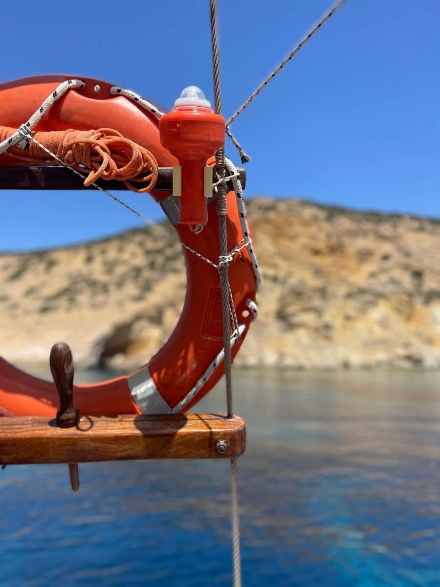 A close-up of a bright orange life preserver and safety equipment on a boat, with a scenic rocky coastline in the background.