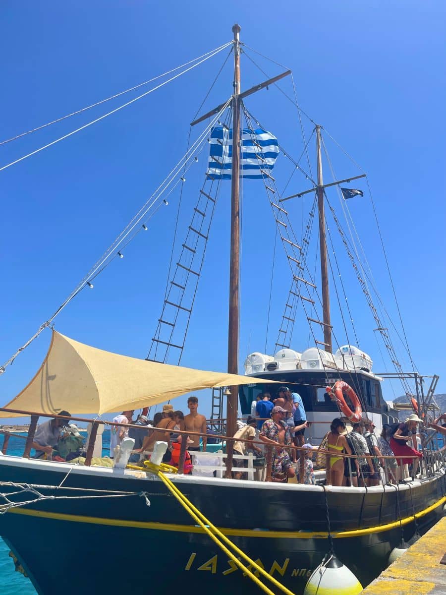 A Greek sailing boat with a blue and white flag docked at a pier, filled with people enjoying the sunny day.