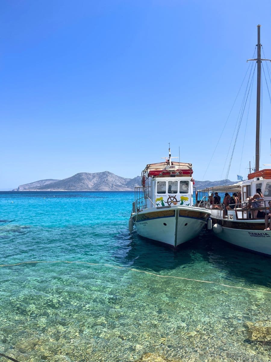 Two boats anchored in crystal-clear shallow waters, with a mountainous island visible in the distance.