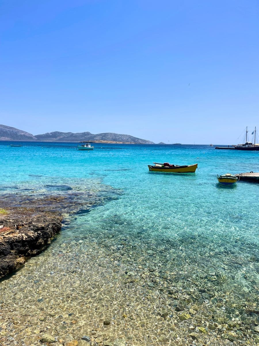 Crystal-clear shallow waters with small boats floating, surrounded by a rocky shoreline and distant islands.