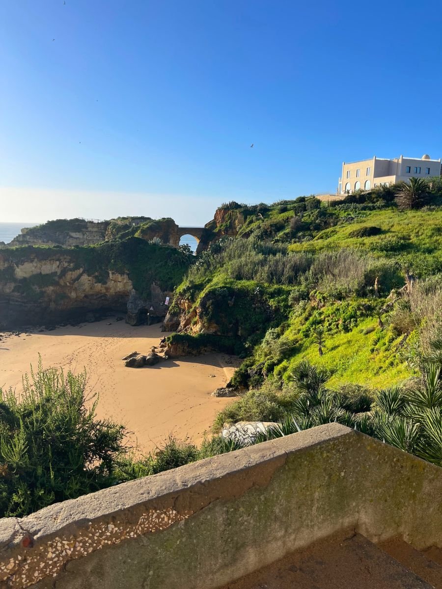View of a quiet beach cove in Lagos, Portugal with lush greenery, rocky cliffs, and a stone arch bridge under clear blue skies.