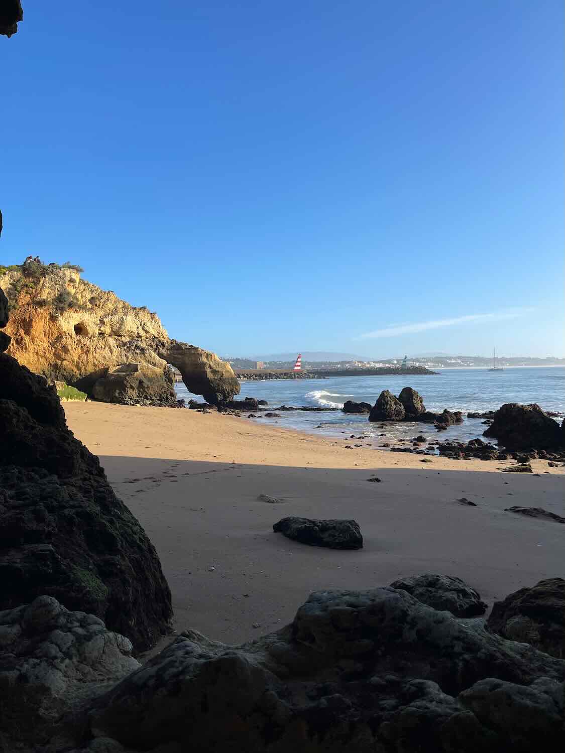 A scenic view of a beach with sandy shore and rocky formations. The sea is calm with small waves, and a distant lighthouse is visible. The sky is clear and blue.
