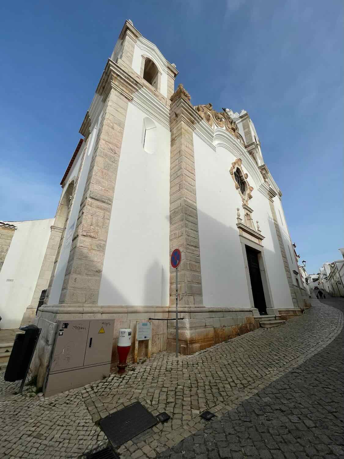A tall white church with intricate stone detailing, viewed from a low angle on a cobblestone street. The sky is clear and blue.