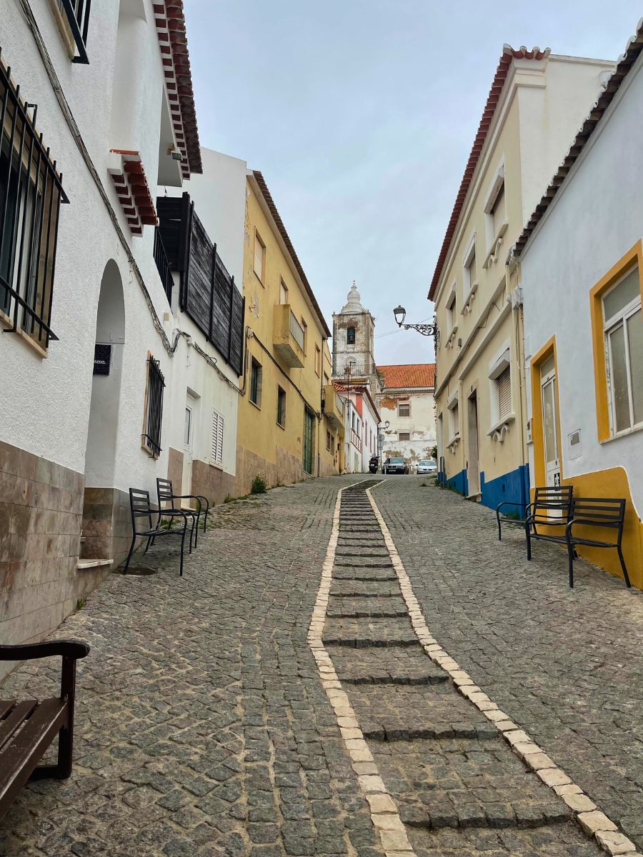 Steep cobblestone street lined with white and yellow buildings in Lagos, with a church tower in the distance