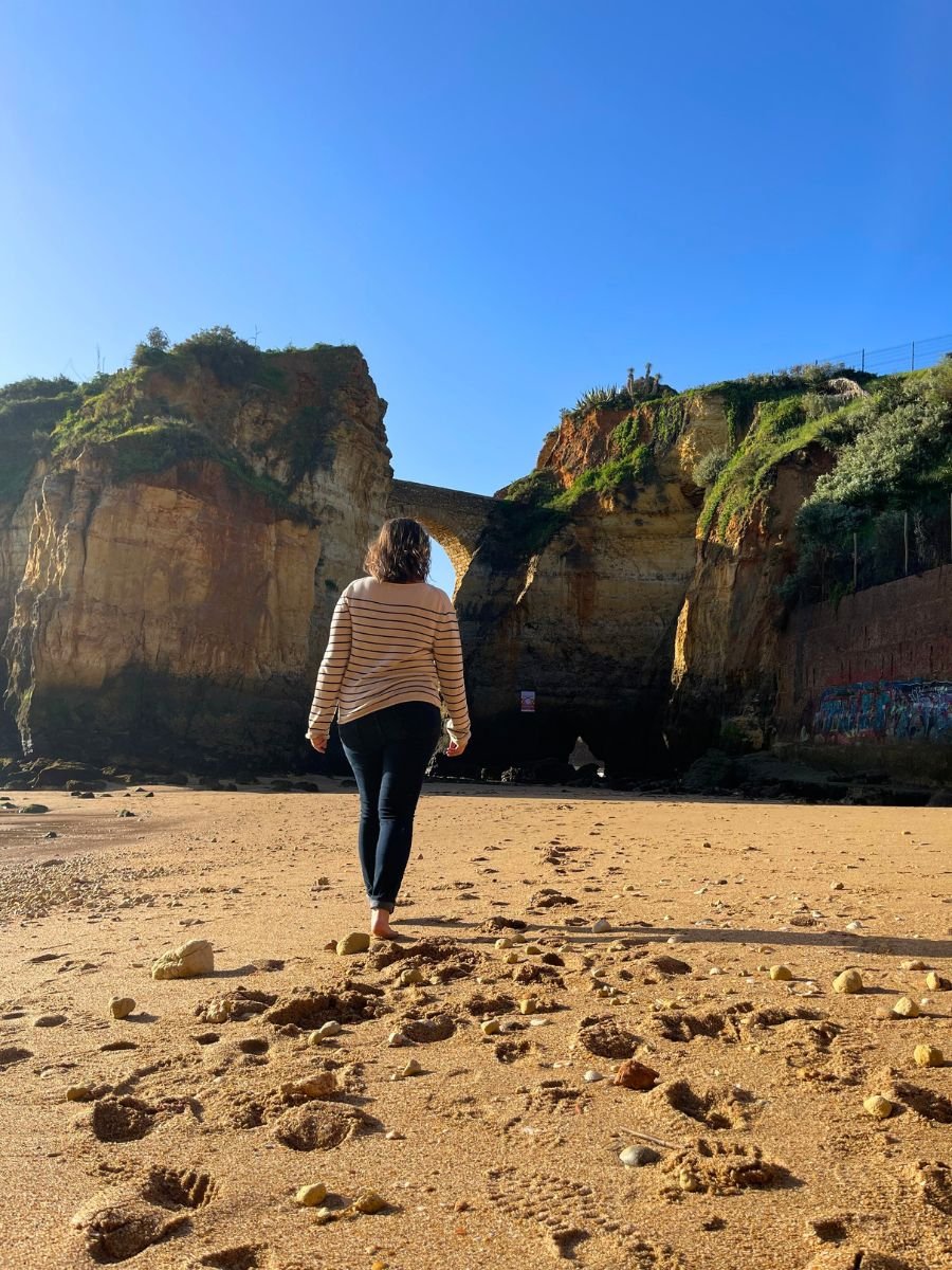 Woman walking barefoot toward a rock arch between cliffs on a beach in Lagos, Portugal.