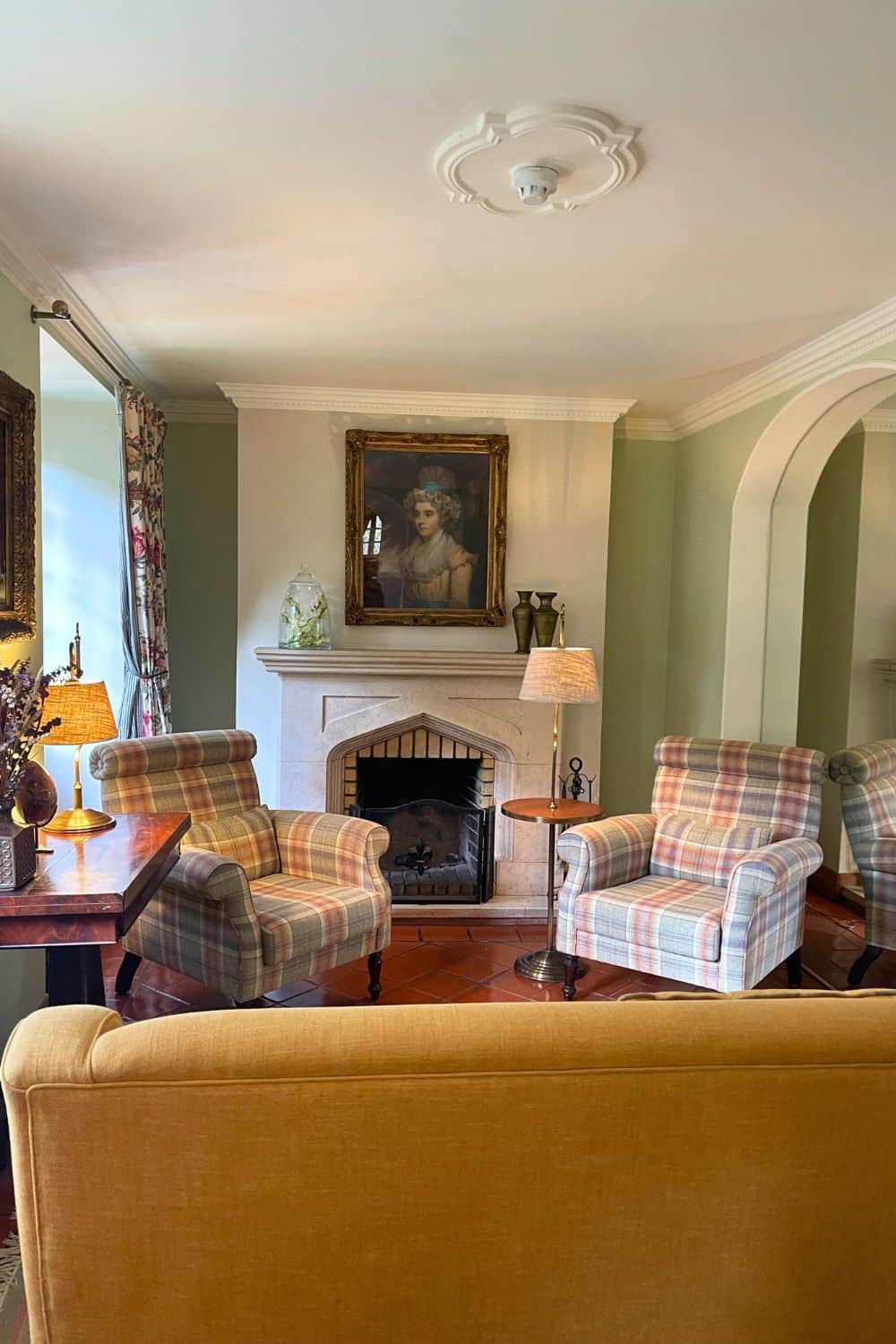 A cozy hotel lounge with plaid armchairs, a fireplace, a classic portrait above the mantle, and soft lighting, creating a warm, inviting atmosphere.