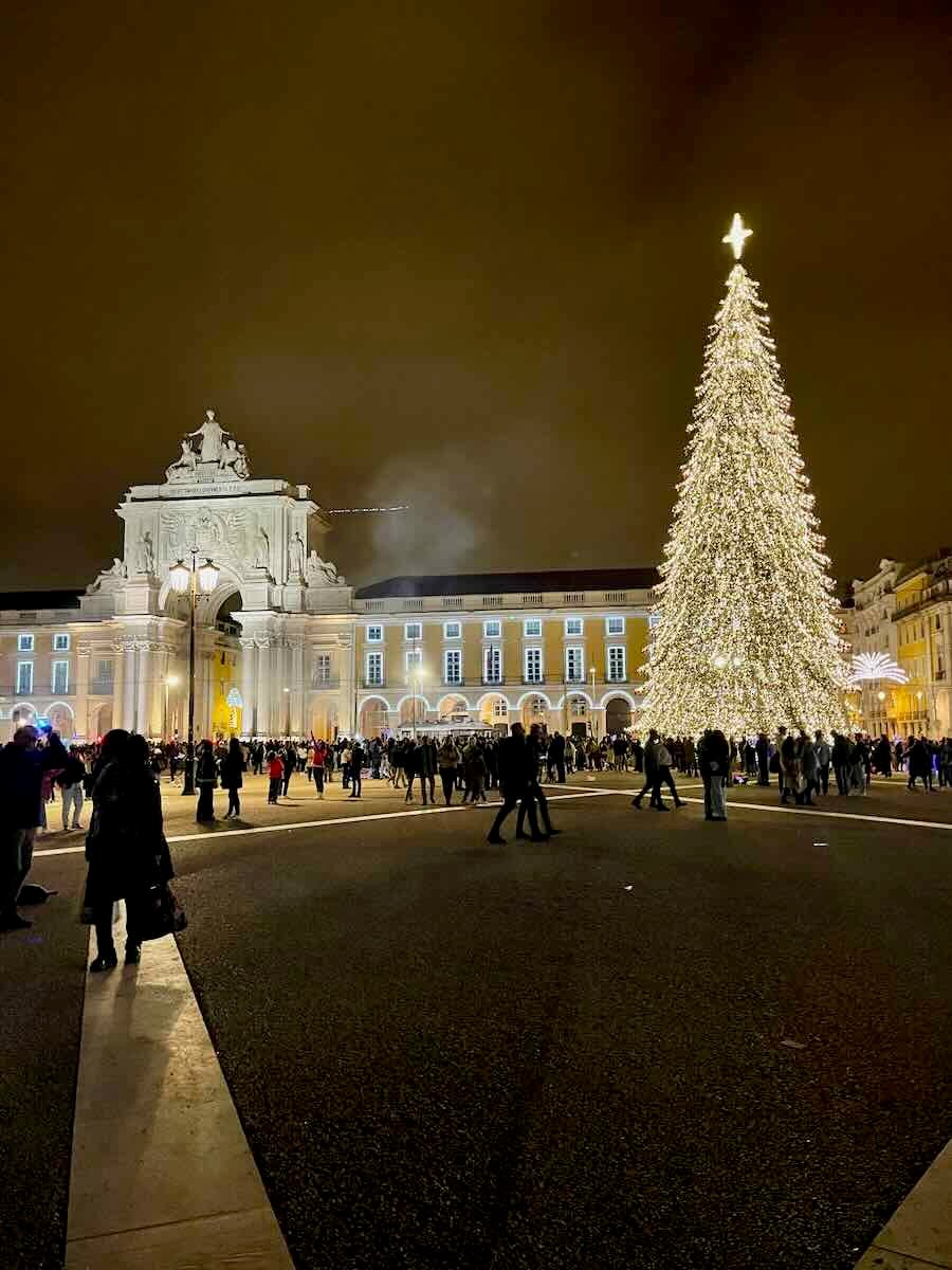 A nighttime scene of a large Christmas tree beautifully lit up in the Praça do Comércio, Lisbon, Portugal, with the historic Arco da Rua Augusta and a crowd of people enjoying the festive atmosphere.